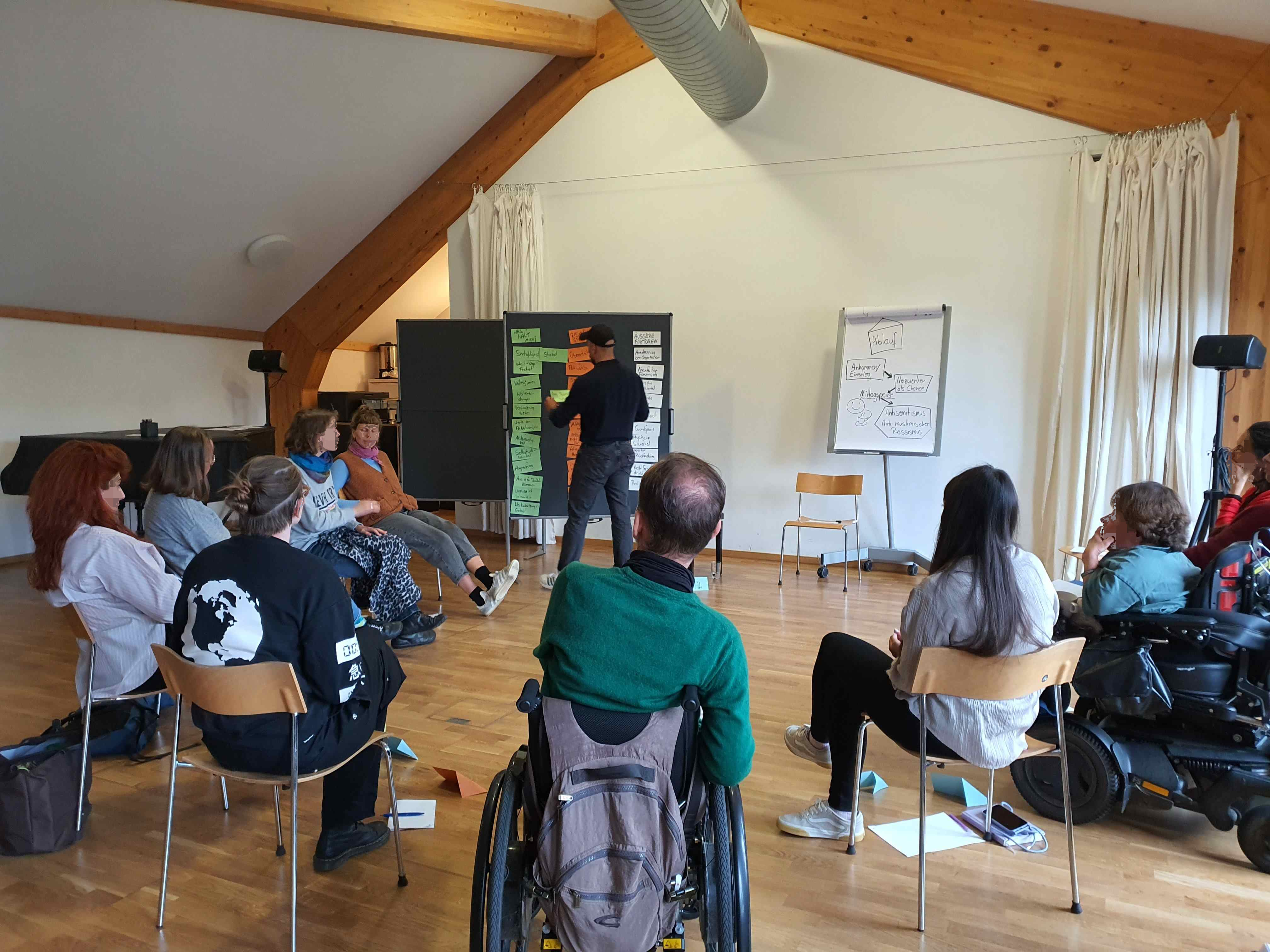 Man sieht mehrere Personen von hinten, die in einem Stuhlkreis sitzen. zwei Personen sitzen im Rollstuhl. Eine Person steht mit dem Rücken zu uns und pinnt gerade ein Moderationskärtchen an eine Pinnwand, an der auch schon andere Moderationskärtchen hängen.