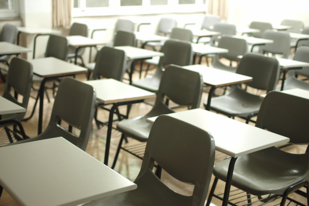 Ein leeres Klassenzimmer. Schmale weiße Tische und dunkelgraue Stühle stehen in Reihen. Sonnenlicht fällt in den Raum.